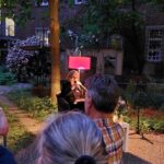 Eine Frau sitzt in einem Sessel und liest während einer Abendveranstaltung im Freien aus einem Buch, das von einer rosa Lampe beleuchtet wird. Die Zuhörer sitzen vor ihr, umgeben von Grünpflanzen und einem Backsteingebäude.