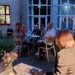 Eine Gruppe von fünf Personen sitzt am Abend draußen an einem Tisch und blickt auf eine Frau im Vordergrund. Die Umgebung scheint ein Innenhof zu sein, mit Blumen und Kerzen auf einem Tisch in der Nähe und einem Gebäude im Hintergrund.