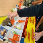 Mehrere Kinder interagieren mit einer Tischplatte, die mit verschiedenen Gegenständen und roten Karten gefüllt ist, darunter ein falsches Tierfell, Sonnencreme und Postkarten, möglicherweise für eine pädagogische oder interaktive Aktivität.