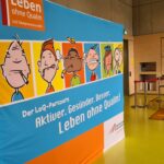 Ein farbenfrohes Display mit Cartoon-Gesichtern und deutschem Text: Leben ohne Qualm und Aktiver. Gesünder. Besser. Leben ohne Qualm! steht in einem Raum mit hellen Holzwänden und gelbem Boden.