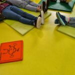 Drei Personen liegen auf grünen Matten auf einem gelben Boden, ihre Beine sind sichtbar und ihre Köpfe sind nicht zu sehen. Zwei tragen Papierkronen an den Füßen. Im Hintergrund sind bunte Matten und ein Spielplatz für Kinder zu sehen.