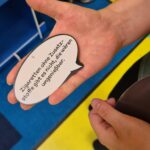 Eine Person hält eine Sprechblase aus Pappe, auf der auf Deutsch steht: Zigaretten ohne Zusatzstoffe gibt es nicht, sie wären ungenießbar. Zwei Personen stehen in der Nähe auf einem blau-gelben Boden.