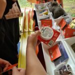 Mehrere Personen stehen um einen Tisch mit verschiedenen Gegenständen, darunter Snacks, rote Karten und ein Plüschtier. Eine Person schaut sich ein rundes Etikett genau an, während andere die Auswahl durchstöbern. Draußen vor dem Fenster sind Bäume zu sehen.