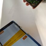 Eine Person hält ein Smartphone mit russischem Text über ein Tablet auf einem Ständer, das eine Website mit einer gelben Menüleiste und deutschem Text auf weißem Hintergrund zeigt. Beide Geräte stehen auf einer weißen Fläche.
