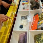 Die Hände einer Gruppe von Personen interagieren mit Gegenständen in einer gelb-weißen Vitrine, die verschiedene Objekte enthält, darunter Pinsel, Pakete, Karten und Werkzeuge.