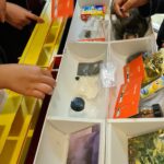 Vier Personen greifen in geordnete weiße Tabletts, die mit verschiedenen drogenspezifischen Gegenständen gefüllt sind, darunter Beutel mit Pulver, Pillen, Blättchen und Aufklärungskarten, die auf einem gelben Tisch bei einer Informationsausstellung oder einem Workshop ausgestellt sind.