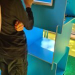 Ein Tablet-Bildschirm zeigt eine deutsche Quizfrage über Stoffe im Tabakrauch, die Optionen wie Nikotin, radioaktives Plutonium, Kohlenmonoxid, Kakao, Blausäure, Formaldehyd, Lakritz und Teer aufführt. Es ist eine Abbildung einer brennenden Zigarette zu sehen.