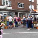 Eine Gruppe von Menschen versammelt sich während eines Marktes im Freien um die Pflanzenstände in einer Straße der Stadt. Einige stöbern in orangefarbenen Kisten nach Pflanzen, während andere sich unterhalten. Im Hintergrund sind ein Backsteingebäude und ein Schaufenster zu sehen.