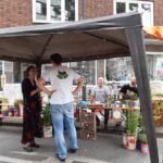 Eine Gruppe von Menschen steht und sitzt unter einem Vordach an einem Marktstand im Freien, umgeben von Topfpflanzen und handgefertigten Waren. Ein Mann in einem weißen Hemd mit einem Pflanzenlogo unterhält sich mit einer Frau in einem langen Rock. Im Hintergrund sind Backsteingebäude zu sehen.