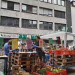 Menschen versammeln sich an einem Marktstand aus Holzpaletten, der von Blumenkisten und bunten Schildern umgeben ist. Im Hintergrund ist ein Gebäude mit vielen Fenstern zu sehen, und über der Menge hängen grüne Wimpel.