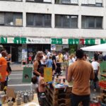 Menschen besuchen einen lebhaften Straßenmarkt mit Ständen, die Pflanzen und Kunsthandwerk verkaufen. Bunte Banner hängen über dem Markt, und eine Menschenmenge schlendert zwischen den Tischen vor einem Gebäude mit großen Fenstern.