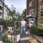Ein Mann steht auf einer Straße in der Nähe von Topfpflanzen und bunten Blumenarrangements auf einem Markt im Freien, mit Menschen und Gebäuden im Hintergrund.