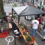 Eine Straßenfest-Szene mit Menschen, die sich um ein überdachtes Zelt versammelt haben, in dem Topfpflanzen auf Bänken ausgestellt sind. Ein Mann sitzt lächelnd da, während andere in der Nähe stehen. Bunte Plakate und Pflanzentöpfe schmücken den Aufbau auf einem städtischen Bürgersteig.