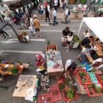 Ein belebter Straßenmarkt mit Menschen, die spazieren gehen und sich versammeln. Auf den Tischen stehen Topfpflanzen, Kunsthandwerk und bunte Gegenstände. Zelte und Dekorationen säumen die Straße, und im Hintergrund ist ein Geschäft mit einer grünen Markise zu sehen.