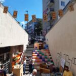 Eine farbenfrohe Veranstaltung im Freien findet auf einer mit bunten Kacheln geschmückten Treppe statt. Die Leute gehen spazieren, stöbern in den Tischen mit den Waren, und über der Szene steht in Pappbuchstaben "QUERBEET". Es ist sonnig und der Himmel klar.