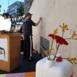 Eine Person malt auf einer großen Außenwand bei einer Straßenveranstaltung. Im Vordergrund steht ein weißer Container mit roten Blumen auf einem bunten Boden. Im Hintergrund sind Wimpel und Menschen zu sehen.