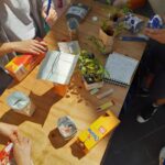 Vier Personen arbeiten an einem Holztisch und stellen Pflanzgefäße aus recycelten Kartons her. Auf dem Tisch liegen Pflanzen, Stifte, ein Feuerzeug, ein Notizbuch mit Skizzen und leere Kartons verstreut.