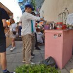 Menschen versammeln sich auf einem Outdoor-Markt oder einer Pop-up-Veranstaltung. Ein Mann mit weißem Hemd und blauer Mütze bereitet an einer rosafarbenen Theke ein Getränk zu, während andere stöbern oder herumlaufen. Im Vordergrund stehen grüne Topfpflanzen.
