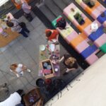 Draufsicht auf Menschen, die an Tischen im Freien, in der Nähe von bunt gestreiften Bänken und Treppen, an Kunsthandwerk arbeiten. Kunstzubehör und Topfpflanzen sind zu sehen, und mehrere Personen sind mit kreativen Tätigkeiten beschäftigt.