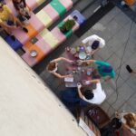 Eine Gruppe von Menschen versammelt sich um einen Tisch mit Malutensilien und malt gemeinsam im Freien an einer bunt gestreiften Treppe, während andere in der Nähe sitzen und zusehen. Die Szene wird von oben betrachtet.
