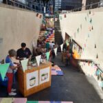Bunte Blumen in recycelten, regenbogenfarbenen Blechdosen sind auf orangefarbenen Regalen angeordnet. Eine schwarz-weiße Comicseite ist teilweise hinter den Dosen auf dem obersten Regal sichtbar. Helles Sonnenlicht erhellt die Szene.