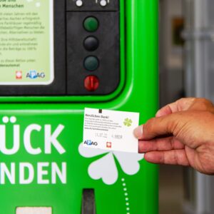 Eine Hand hält ein gedrucktes Ticket vor einem hellgrünen Rückgabeautomaten mit deutschem Text und verschiedenen Tasten, Etiketten und Schlitzen, die auf dem Automaten zu sehen sind.