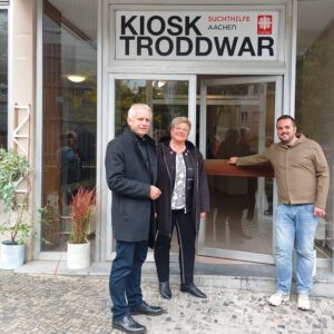 Drei Personen stehen lächelnd vor einem Hauseingang mit einem Schild mit der Aufschrift Kiosk Troddwar Suchthilfe Aachen. Zwei Männer und eine Frau posieren auf einem kopfsteingepflasterten Bürgersteig neben Topfpflanzen.