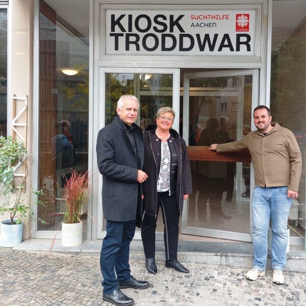 Drei Personen stehen lächelnd vor einem Hauseingang mit einem Schild mit der Aufschrift Kiosk Troddwar Suchthilfe Aachen. Zwei Männer und eine Frau posieren auf einem kopfsteingepflasterten Bürgersteig neben Topfpflanzen.