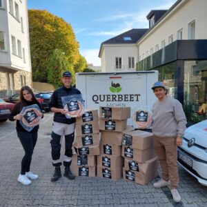 Drei Personen stehen vor einem Stapel Pappkartons und einem Schild von Querbeet Aachen, jede hält ein verpacktes Lebensmittel in der Hand, draußen auf einer gepflasterten Fläche, umgeben von Gebäuden und geparkten Autos.