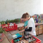 Eine Frau mit Brille und weißem Hemd arbeitet an einem Holztisch, der mit Farbsprühdosen, Schachteln und bunten Töpfen bedeckt ist, mit weiteren bemalten Töpfen und Pflanzen im Hintergrund.