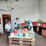 Drei Personen in weißen Hemden und blauen Handschuhen bemalen bunte zylindrische Objekte auf einer Holzpalette in einer Industriehalle mit weißen Wänden und vereinzelten Pflanzen.