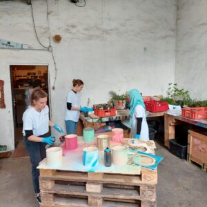 Drei Personen in weißen Hemden und blauen Handschuhen bemalen bunte zylindrische Objekte auf einer Holzpalette in einer Industriehalle mit weißen Wänden und vereinzelten Pflanzen.