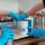 Drei Personen mit blauen Handschuhen arbeiten zusammen, um eine Blechdose mit einem dekorativen Etikett auf einem Tisch aus Holzpaletten zu besprühen und zu halten, während im Hintergrund weitere Dosen und bunte Materialien zu sehen sind.