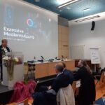 Eine Frau spricht an einem Podium in einem Konferenzraum. Auf einem Bildschirm hinter ihr ist der Titel Exzessive Mediennutzung bei Kindern und Jugendlichen zu sehen. Mehrere Personen sitzen und hören zu, und rechts ist ein Flipchart zu sehen.