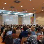 Eine große Gruppe von Menschen sitzt in einem Vortragssaal und hört einem Redner zu, der im vorderen Bereich einen Vortrag hält. Auf einer Leinwand erscheint der deutsche Satz Exzessive Mediennutzung. Die Zuhörer blicken aufmerksam nach vorne.