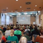Ein großes Publikum sitzt in einem modernen Hörsaal und blickt auf einen Vortragenden an der Stirnseite. Der Vortragende steht neben einer Leinwand, auf der die Worte "Exzessive Mediennutzung" zu lesen sind, was auf einen Vortrag über exzessive Mediennutzung hinweist.