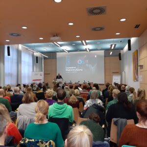 Ein großes Publikum sitzt in einem modernen Hörsaal und blickt auf einen Vortragenden an der Stirnseite. Der Vortragende steht neben einer Leinwand, auf der die Worte "Exzessive Mediennutzung" zu lesen sind, was auf einen Vortrag über exzessive Mediennutzung hinweist.