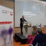 Ein Mann im Anzug spricht an einem Podium in einem Konferenzraum. Hinter ihm ist ein Bildschirm mit der Aufschrift Exzessive Mediennutzung und den Logos von Caritas und Suchthilfe Aachen zu sehen. Im Vordergrund sind Blumen und ein RESERVIERT-Schild zu sehen.
