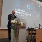 Ein Mann steht an einem Podium und hält einen Vortrag mit dem Titel "Exzessive Mediennutzung bei Kindern und Jugendlichen" in einem Konferenzraum, mit Blumen auf dem Podium und einer Projektionsfläche im Hintergrund.