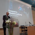 Ein Mann im Anzug spricht an einem Podium in einem Konferenzraum. Hinter ihm ist eine Präsentationsfolie mit der Überschrift Exzessive Mediennutzung bei Kindern und Jugendlichen" zu sehen. Auf dem Tisch neben ihm stehen Blumen und Wasserflaschen.
