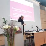 Ein Mann steht an einem Podium in einem Konferenzraum und hält einen Vortrag mit dem Titel Prävention von Mediensucht. Im Vordergrund sind Blumen arrangiert, und auf dem Tisch neben ihm stehen Stühle, ein Mikrofon und eine Wasserflasche.