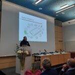 Ein Mann hält einen Vortrag auf einem Podium in einem Konferenzraum. Auf einem Bildschirm ist eine Folie mit dem Titel "Digitale Alleskönner" und Icons von digitalen Geräten zu sehen. Mehrere Personen sitzen und schauen zu. Vor dem Podium sind Blumen arrangiert.