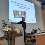 Ein Mann steht an einem Podium in einem Vortragsraum und präsentiert eine Folie mit dem Titel Faszination und dunkle Aspekte - Muster in Spielen, wobei ein animiertes Bild auf dem Bildschirm angezeigt wird. Auf dem Pult stehen Blumen und eine Flasche Wasser.