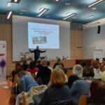 Ein Redner steht an einem Podium und hält einen Vortrag zum Thema "Exzessive Mediennutzung" vor einem Publikum in einem Konferenzraum. Eine Leinwand zeigt das Thema, links ist ein Banner der Suchthilfe Aachen zu sehen.