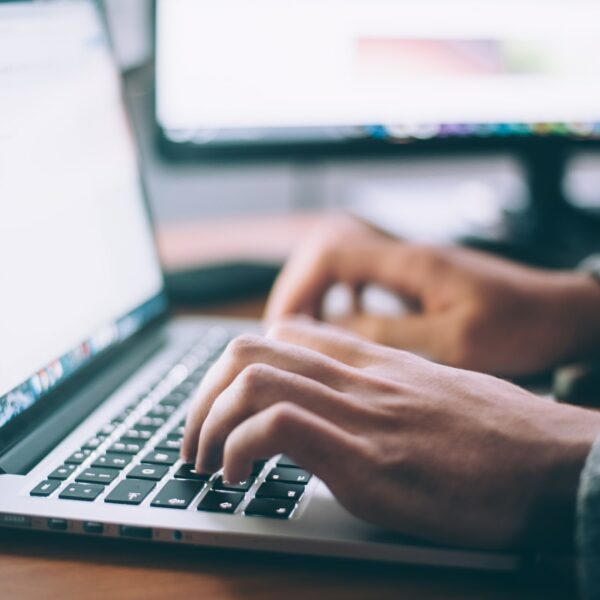 Eine Person tippt auf einer Laptop-Tastatur an einem Schreibtisch, im Hintergrund ist ein weiterer Computermonitor zu sehen. Die Szene deutet auf eine Arbeits- oder Studienumgebung hin.