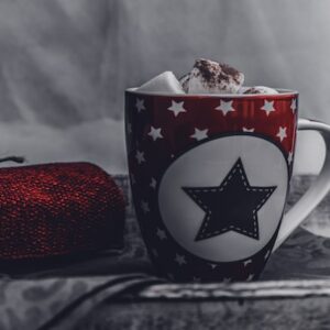 Ein roter Becher mit einem schwarzen Stern und weißen Sternen darauf, gefüllt mit heißer Schokolade und Marshmallows, steht neben einem zusammengerollten roten Strickschal auf einer dunklen Stoffoberfläche. Der Hintergrund ist sanft verschwommen.