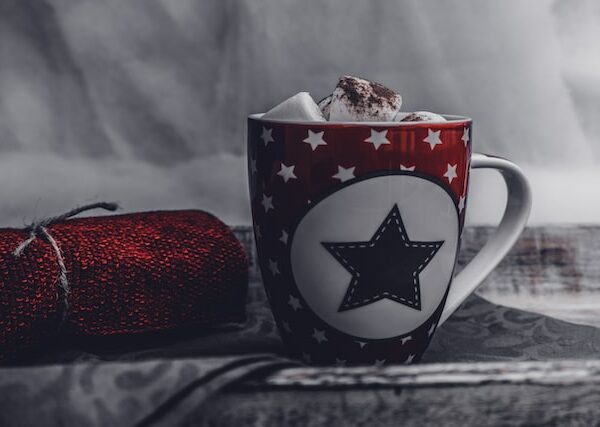 Ein roter Becher mit einem schwarzen Stern und weißen Sternen darauf, gefüllt mit heißer Schokolade und Marshmallows, steht neben einem zusammengerollten roten Strickschal auf einer dunklen Stoffoberfläche. Der Hintergrund ist sanft verschwommen.