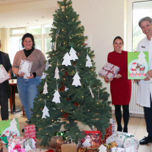 Vier Erwachsene stehen in einem Haus neben einem geschmückten Weihnachtsbaum und halten jeweils eingepackte Geschenke in der Hand. Zahlreiche Geschenke liegen unter dem Baum, und an den Zweigen hängt weißer Papierschmuck. Die Umgebung wirkt festlich und fröhlich.