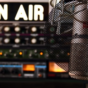Nahaufnahme eines Studiomikrofons vor einem Poppfilter, mit einem beleuchteten ON AIR-Schild und unscharfem Audio-Equipment im Hintergrund.