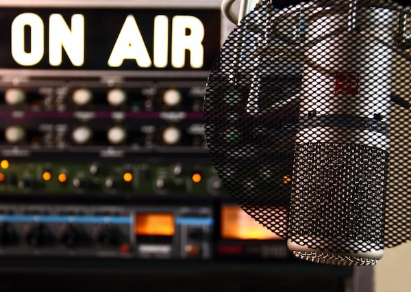 Nahaufnahme eines Studiomikrofons vor einem Poppfilter, mit einem beleuchteten ON AIR-Schild und unscharfem Audio-Equipment im Hintergrund.